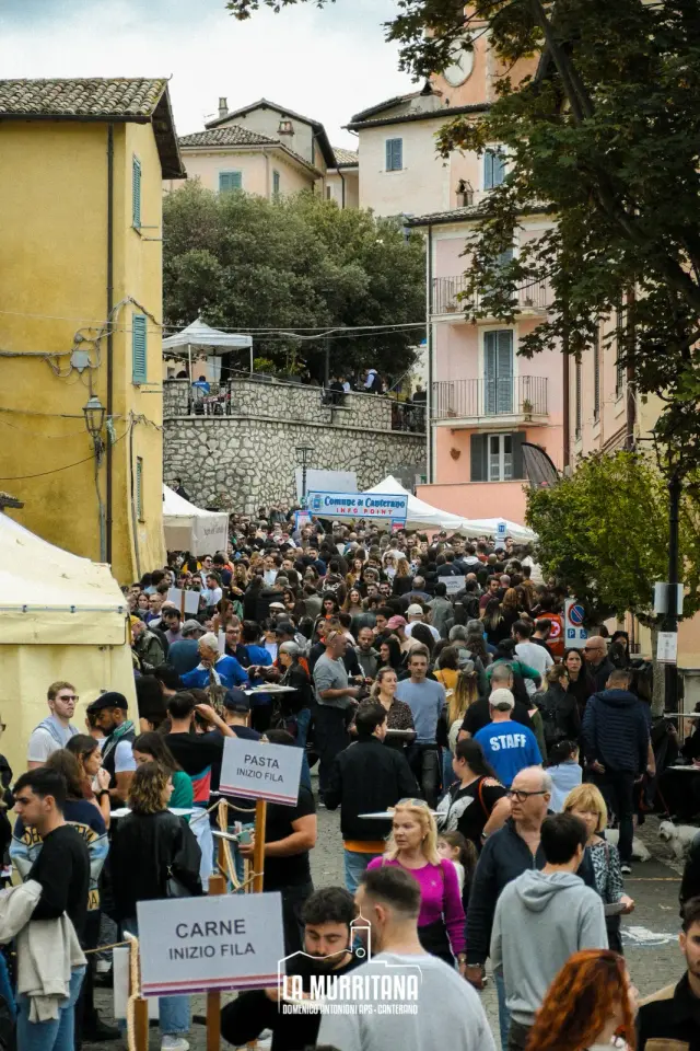 Vicolo del paese affollato di visitatori con cartelli "Pasta inizio fila" e "Carne inizio fila"